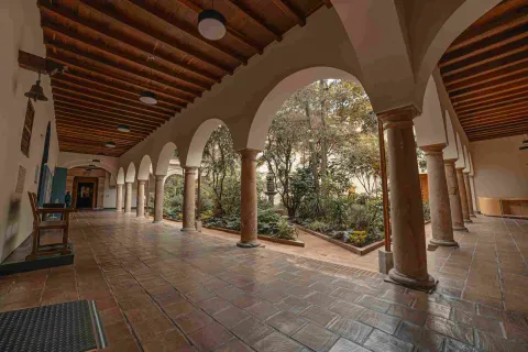 Un primer piso en el que hay un largo pasillo en ángulo con unas columnas que forman arcos y sostienen un segundo piso. Al fondo se observa un patio con un jardín.