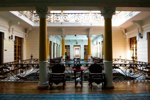 Interior view of an old building showing beautiful and elegant stone walls, bronze frames, and white and yellow columns. A waxed wooden floor is visible.