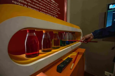 Una exposición de botellas de vidrio con un líquido de color específico en cada una. Hay una mano que parece estar señalando algo.