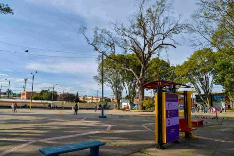 Una amplia plaza pública en un día despejado, con un ambiente tranquilo y cotidiano. El espacio está pavimentado con grandes losas de concreto, marcadas por líneas diagonales de color rosado pálido que crean un diseño geométrico en el suelo. En el primer plano, a la derecha, destaca una estructura informativa de color amarillo y morado con un techo rojo, acompañada de un banco de madera rojiza. A la izquierda, se observa un banco largo y sencillo de color azul intenso.