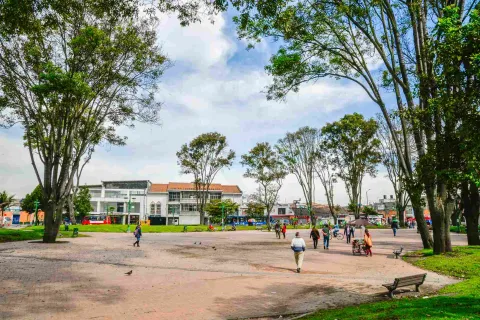 Plano general de la plazoleta central donde hay varios árboles altos y personas que caminan alrededor.