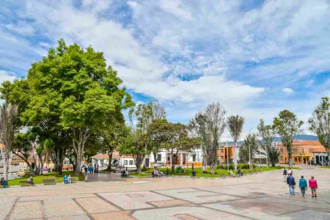 Una plaza de pueblo con edificios coloridos y personas disfrutando del día. En la imagen, se ve una amplia plaza pavimentada con adoquines de color claro. 