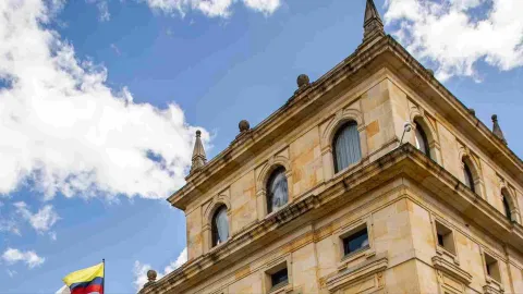 Primer plano de la parte alta de la edificación de piedra, en la que se observan algunas figuras en forma de conos y esferas. A un lado hay una bandera de Colombia en un cielo azul.