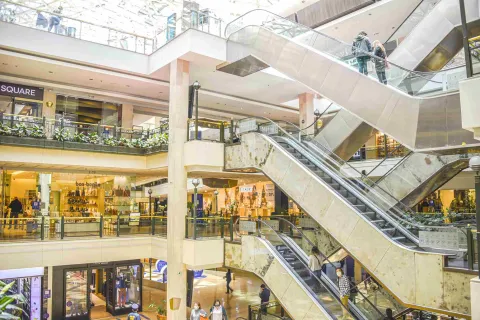 Interior de centro comercial en el que sobresalen escaleras eléctricas, locales comerciales en varios pisos y algunas personas alrededor.