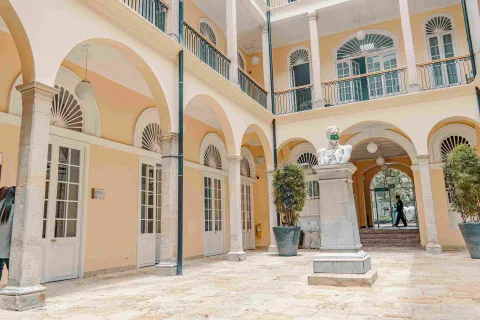  Primer plano del interior del palacio en una especie de patio en el que se observa una estructura de dos pisos color crema, el segundo con bellos balcones y en el primero varias columnas en forma de arco. En el centro del lugar hay una estatua, parece que de un héroe patrio.