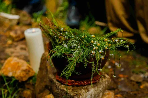 Un tazón de cerámica lleno de flores y plantas está sobre una roca, con una vela blanca cerca.