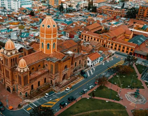 Panorámica de una iglesia vista desde arriba, como con un dron. Sobresalen los techos de ladrillo y una torre alta en forma ovalada. Se observa un parque al frente de la iglesia.