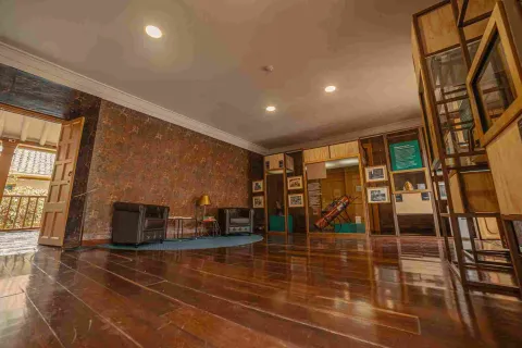 Salón interior con un piso en madera brillante y en el que se observan algunos estantes en madera con diversos artículos, entre ellos un extintor gigante. En el salón hay dos poltronas de cuero, separadas por dos mesas pequeñas; en una de ellas hay una lámpara metálica dorada.