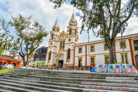 Vista inspiradora de una iglesia histórica con dos campanarios en Bogotá, rodeada de frondosos árboles y vibrante arte callejero en las escaleras, que simboliza el patrimonio cultural y el espíritu comunitario.