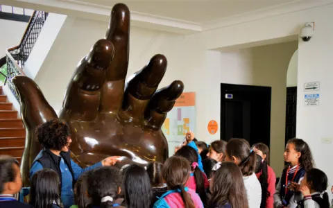 A giant, brown, hand-shaped sculpture surrounded by children and an adult woman who appears to be a guide or teacher.