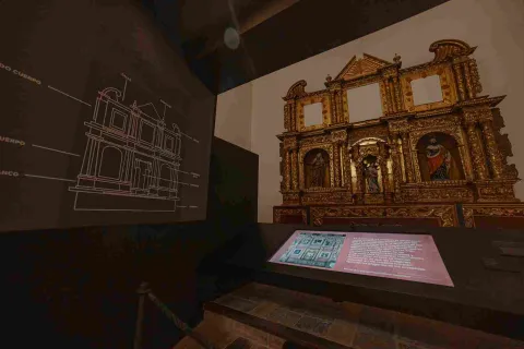 Un pedazo grande de retablo dorado está exhibido en una pared. Hay tres imágenes de santos que lo acompañan. Hay una foto de un plano de una casa que acompaña la escena.