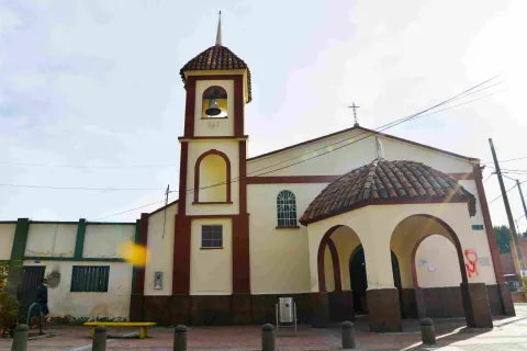  Una iglesia de estilo colonial con una torre campanario prominente. La estructura principal está pintada en tonos crema claro con detalles en rojo ladrillo, creando un contraste visual llamativo. El techo es de tejas rojas, y sobre el edificio principal se alza una cruz.