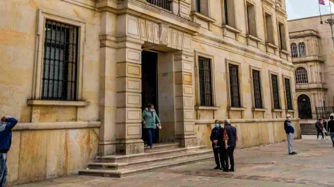 Vista de lado de la fachada del edificio en piedra. Hay una mujer que sale por la puerta principal y unas personas alrededor.