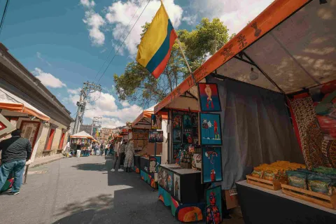 A street lined with vendor stalls displaying handicrafts and paintings, with people browsing in some of them. A Colombian flag hangs prominently on one of the stalls.