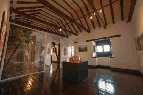Un salón interior en el que sobresale el techo en vigas de madera y, en el centro del piso, sobre una plataforma, una olla sopera grande. Al fondo hay una pequeña ventana por donde entra algo de luz natural.
