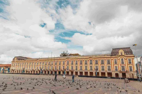 Plano general de la fachada exterior del palacio, vista de frente. Se observan muchas puertas en piedra en el primer piso y muchas ventanas en el segundo. Al frente se ve la Plaza de Bolívar con algunas personas y varias palomas.