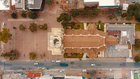 Aerial view of a church with a red-tiled roof, surrounded by a large pedestrian plaza with scattered trees. A road with vehicular traffic is visible to one side, and urban buildings surround it. The composition shows the organization of the public space and the architecture of the area from a top-down perspective.