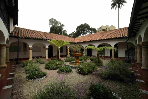 Interior courtyard of a colonial-style building with arched and columned corridors, red tile roofs, and a central garden with plants, ferns, and a small stone fountain.