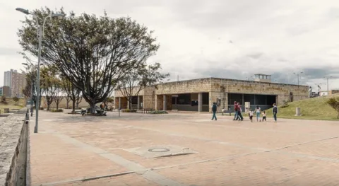 A spacious open-air brick plaza with a low stone building and dark windows in the background. To the left, a large tree provides shade next to benches and lampposts, while several people stroll through the open space under a cloudy urban sky.