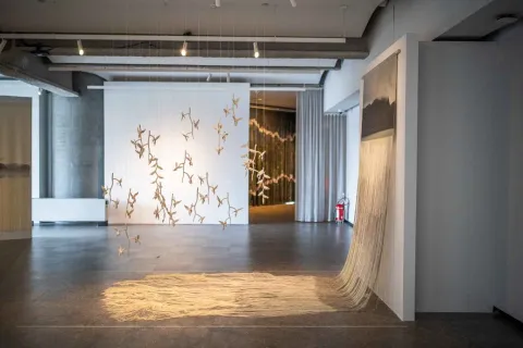 A contemporary art installation in the Bancolombia Art Gallery, where delicate suspended figures hang from the ceiling in front of a white wall, while threads or textile fibers cascade from a structure, forming a kind of canopy on the floor.