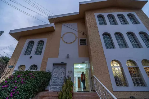 Close-up view of the facade of a two-story house with a beige and white design, characterized by tall windows with arches and metal grilles. In the center, a staircase leads to the main entrance, where a person stands in the illuminated doorway.