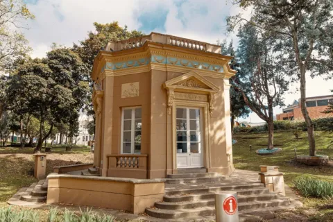 Pequeño quiosco de estilo clásico en medio de un parque, con fachada en tonos beige y detalles ornamentales en relieve y molduras decorativas en azul y dorado. El edificio tiene ventanas y una puerta blanca, además de una escalinata de acceso frontal. Está rodeado de árboles, zonas verdes y senderos, en un entorno tranquilo y luminoso durante el día.