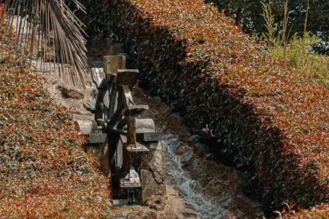 A small wooden waterwheel powered by water in a canal surrounded by shrubs and ornamental vegetation in a park.
