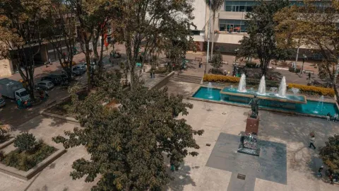 Vista elevada de una plaza urbana con árboles frondosos, senderos peatonales y una fuente rectangular con chorros de agua en funcionamiento. En el centro se observa una estatua sobre pedestal, rodeada de espacios abiertos donde algunas personas caminan o descansan.