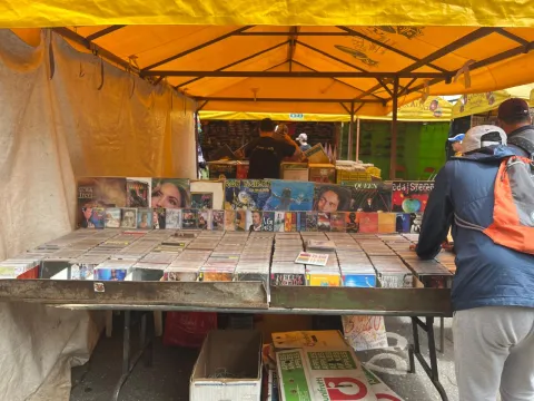Puesto de mercado cubierto con carpa amarilla donde se exhiben discos y CDs organizados en cajas sobre una mesa. En la parte trasera se ven portadas de álbumes de distintos artistas, mientras algunas personas revisan la mercancía.