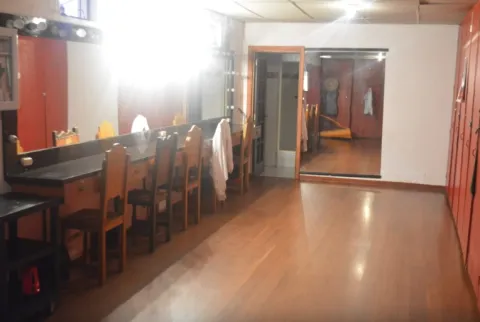 Interior de un camerino de teatro con piso de madera, equipado con un largo tocador con espejos iluminados y varias sillas de madera. A un lado se ven casilleros o armarios rojos, y al fondo un espejo grande refleja parte del espacio.