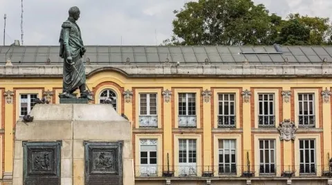 Estatua de un personaje histórico de pie sobre un pedestal, ubicada frente a un edificio de estilo neoclásico con columnas altas y detalles arquitectónicos en piedra. La escultura tiene una pátina verdosa y está rodeada de algunas palomas posadas en la base, en un entorno urbano de carácter histórico y solemne.