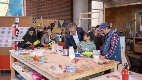Grupo de personas de distintas edades participando en un taller en un espacio tipo laboratorio o biblioteca. Alrededor de una mesa de trabajo con materiales, utensilios y objetos coloridos, un adulto guía a un niño mientras conversan, y otros participantes mezclan ingredientes y realizan actividades manuales.