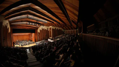  Vista lateral del interior de un gran auditorio durante un concierto. En el escenario de madera se presenta una orquesta sinfónica, mientras el público llena las graderías que se elevan a los lados. El techo alto de madera con vigas geométricas y luces lineales ilumina el recinto, creando una atmósfera cálida para la presentación musical.