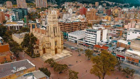 Aerial view of a Neo-Gothic church with a tall tower and ornate facade, located in a large urban plaza. Trees, pedestrians, and buildings of varying heights are visible around it, with a dense urban area and mountains in the background. The scene depicts a busy public space in the city during the day.