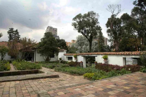 Garden of a colonial house with brick pathways, a small rectangular pond, and white walls with red tiles, surrounded by trees and vegetation, with modern buildings visible in the background.