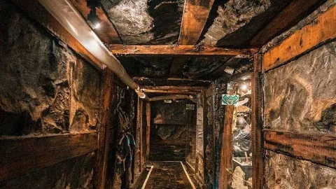 Interior de un túnel minero con paredes de roca y estructuras de madera que sostienen el techo. El pasadizo está iluminado con luces pequeñas y tiene un camino delimitado en el suelo. A un lado se observa una figura que representa a un minero trabajando dentro de la recreación de una mina.