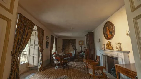  Interior de una sala histórica de la Quinta de Bolívar, con muebles de madera de estilo colonial, una mesa redonda con sillas, cortinas largas junto a ventanas abiertas y una chimenea decorada con relojes y retratos antiguos en la pared. La habitación conserva una ambientación que evoca la vida doméstica del siglo XIX.