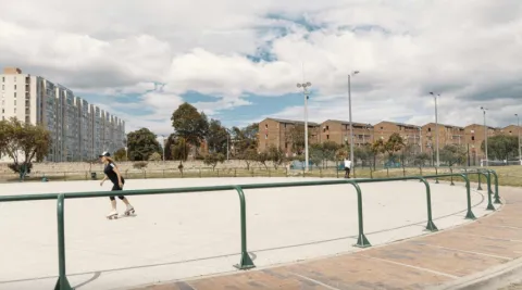 An outdoor skating rink surrounded by green railings, where a person skates on the concrete. In the background, residential buildings, trees, and lampposts are visible under a partly cloudy sky, in a wide-open urban setting.
