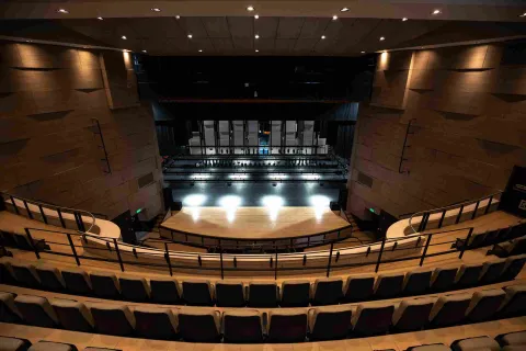 Vista desde las graderías del interior de un teatro moderno con asientos organizados en forma semicircular. Al frente se observa el escenario iluminado y la estructura técnica detrás del telón, con paredes en tonos cálidos y un techo con luces empotradas que iluminan el auditorio.