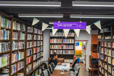  Interior de una biblioteca con estanterías de madera llenas de libros a ambos lados. En el centro hay una mesa larga con varias sillas donde una persona está sentada leyendo o trabajando con un libro. En la parte superior cuelga un letrero morado que dice “Sala general”, acompañado de banderines decorativos y luces fluorescentes en el techo. Al fondo se observan más estanterías y un espacio de lectura.