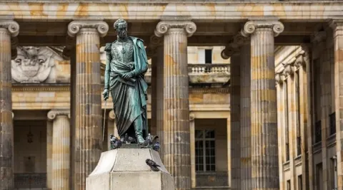 Estatua de un personaje histórico sobre un pedestal de piedra, vista de perfil, frente a un edificio de estilo colonial con fachada amarilla, ventanas simétricas y balcones decorados. En la base de la escultura hay algunas palomas, y al fondo se observan árboles que enmarcan este entorno urbano de carácter histórico.