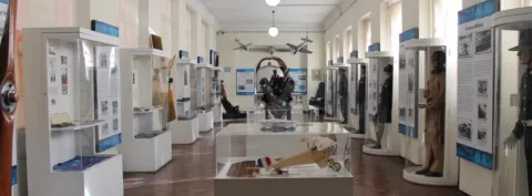 Sala de museo con vitrinas que exhiben objetos históricos militares y de aviación, incluyendo uniformes, maquetas de aviones y paneles informativos dispuestos a lo largo de un pasillo iluminado.