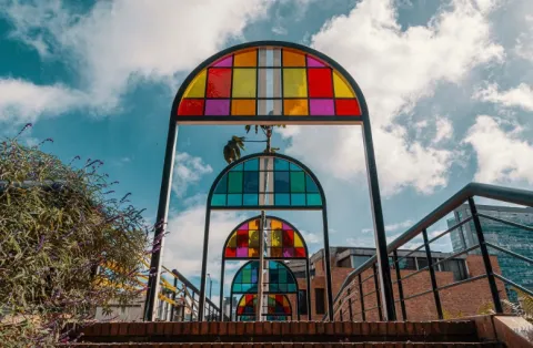 Estructura artística en forma de arcos metálicos con paneles de vidrio de colores, dispuesta en secuencia sobre un sendero. Los vitrales en tonos rojo, amarillo, azul y verde contrastan con el cielo parcialmente nublado al fondo. A los lados se observan barandas, vegetación y edificaciones urbanas, creando un espacio moderno y visualmente llamativo.