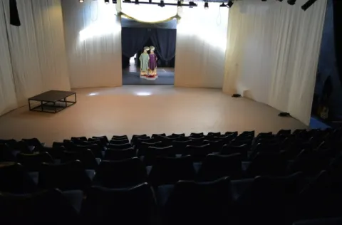 Interior de una sala de teatro con filas de sillas oscuras frente a un escenario sencillo cubierto con cortinas blancas. En el fondo del escenario, a través de una abertura con telones oscuros, se observan figuras o vestuarios coloridos dispuestos como parte de la escenografía.