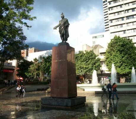 Estatua de bronce de un personaje histórico sobre un pedestal alto en una plaza urbana, con la inscripción “Santander” visible en la base. Alrededor se observan fuentes de agua en funcionamiento, árboles y edificios altos, mientras varias personas caminan por el espacio.