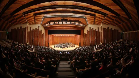 Vista frontal panorámica del interior de un gran auditorio durante un concierto sinfónico. En el escenario de madera se encuentra una orquesta con numerosos músicos e instrumentos, mientras el público llena las graderías y platea a ambos lados. El recinto destaca por su techo curvo de madera con vigas visibles y una iluminación cálida que enmarca el escenario.