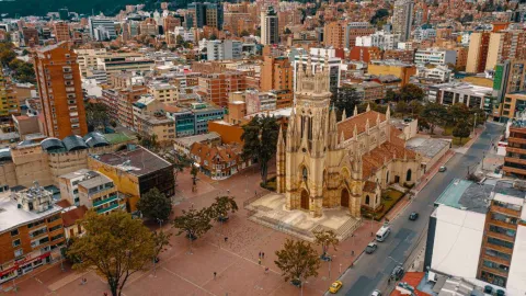 Aerial view of a Neo-Gothic church with a tall central tower and a detailed stone facade, located in the middle of a large pedestrian plaza. Trees, people walking, and roads with vehicular traffic are visible around it, along with numerous residential and commercial buildings that make up a dense urban environment, with mountains visible in the background.