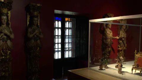 Museum room with red walls displaying antique religious sculptures; to the left are carved figures in the Baroque style, and to the right, a glass display case with small pieces.