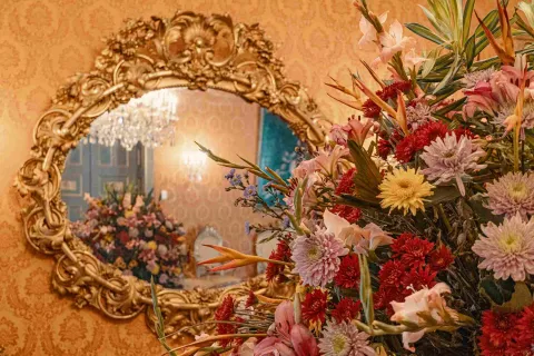 Primer plano de un arreglo floral colorido con flores rojas, rosadas, amarillas y naranjas, dispuesto en un elegante salón de estilo clásico. Al fondo se observa un gran espejo con marco dorado ornamentado que refleja un candelabro de cristal y más flores, creando un ambiente sofisticado y decorativo.
