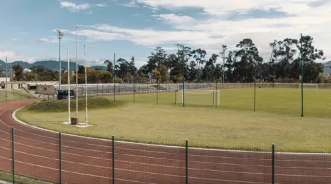 An outdoor sports facility with a red running track surrounding a green grass soccer field, enclosed by a chain-link fence. In the background, bleachers, tall trees, and mountains are visible under a partly cloudy sky.